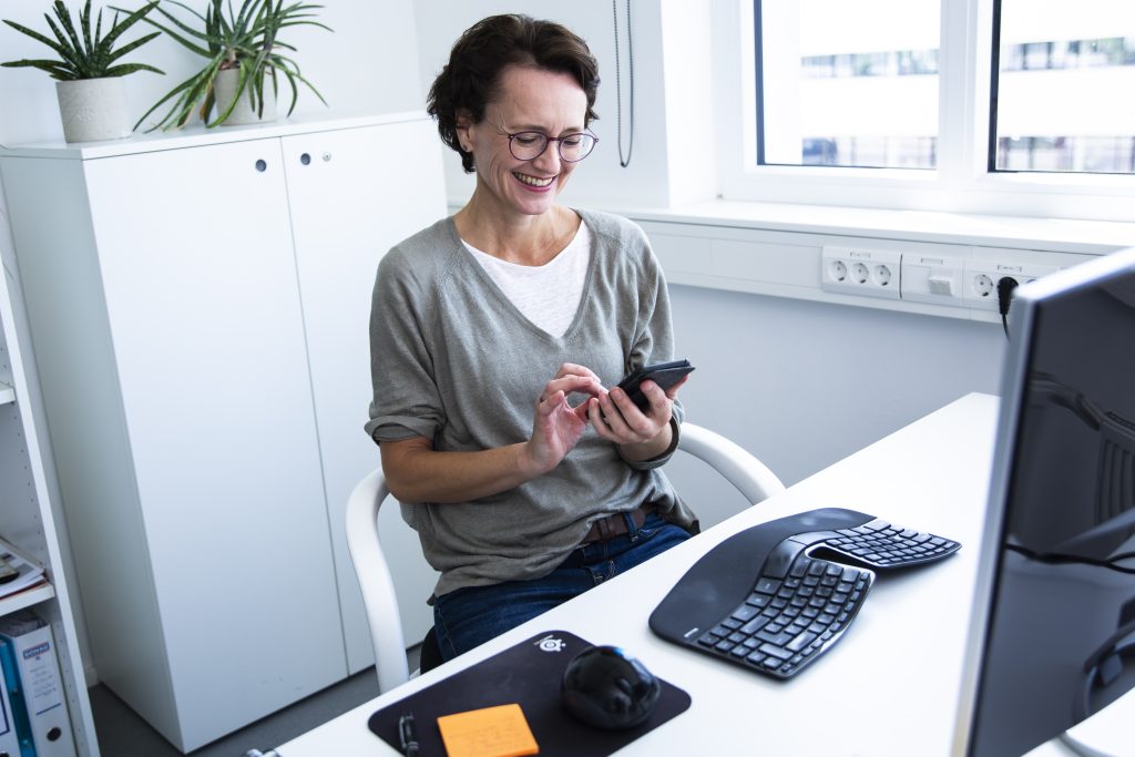 Onine-Business: Eva Dragosits sitzt gutgelaunt am Schreibtisch und schaut aufs Handy