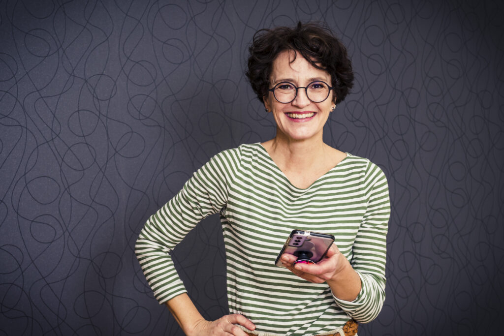 Frau in gestreiftem Shirt hält das Handy in der Hand und strahlt in die Kamera